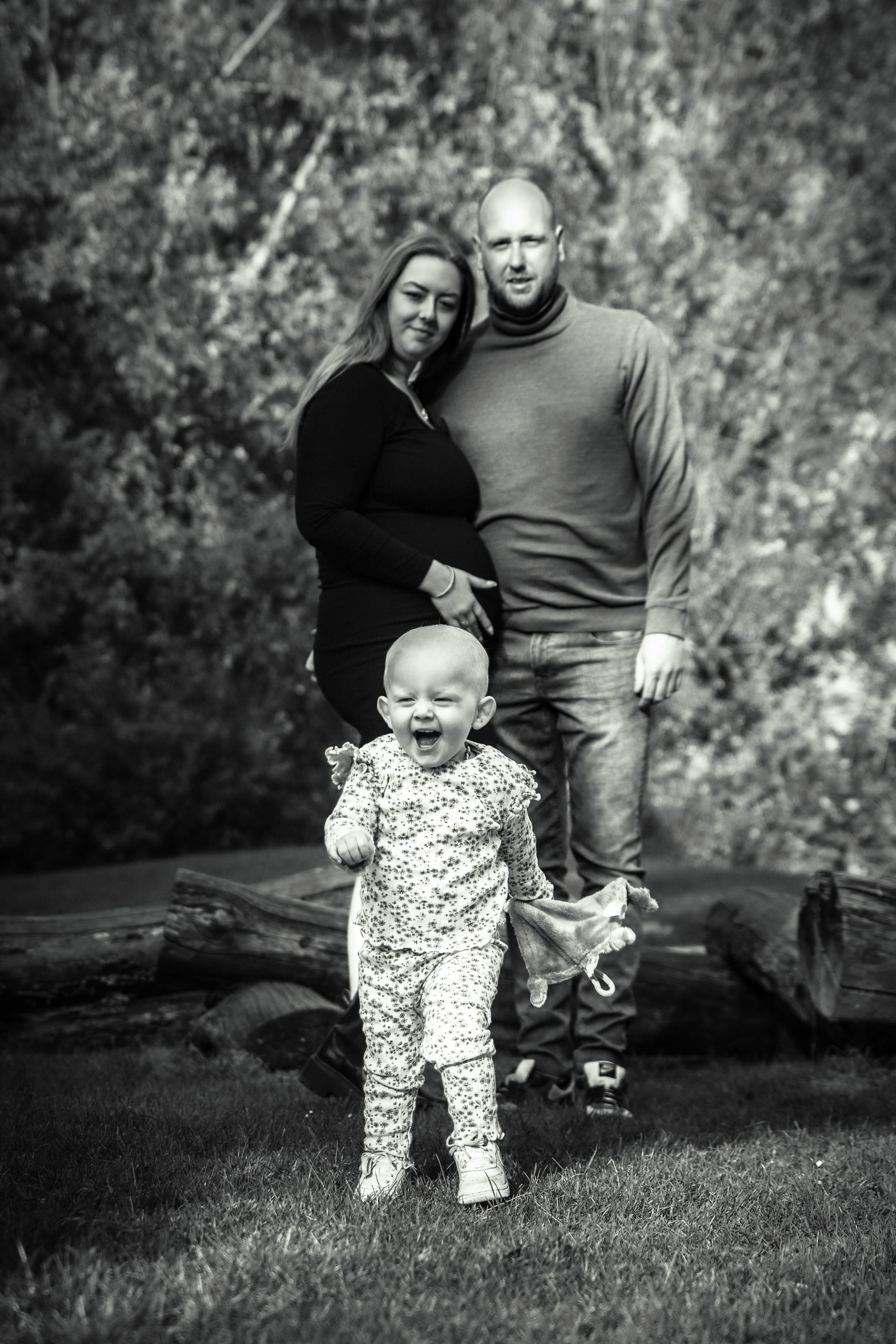 Een gezin van drie poseert buiten op een boomstam, met een lachende peuter vooraan en twee volwassenen erachter. Genderreveal