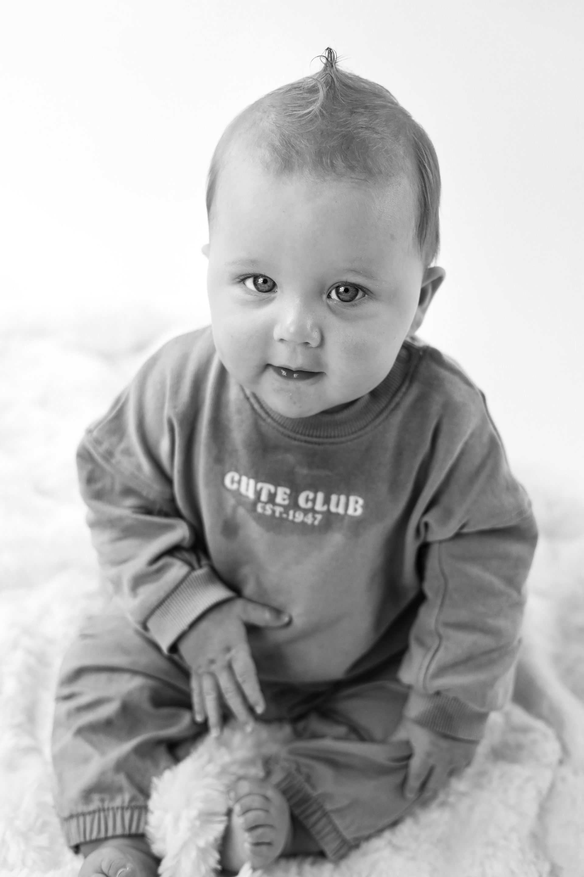 De baby zit op een dekentje, draagt een trui en een broek, en kijkt met een lichte glimlach naar de camera. Kinderfotoshoot
