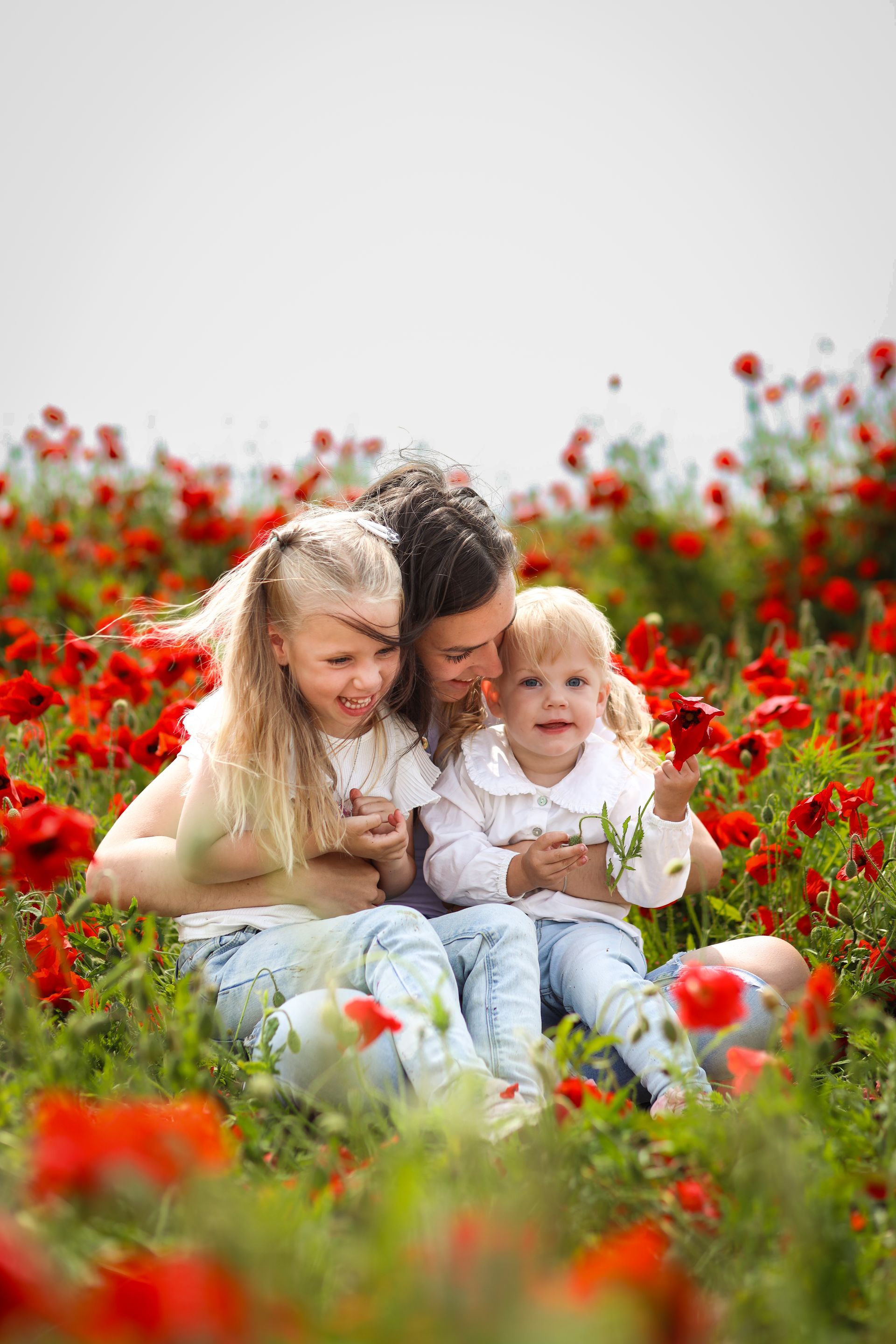 Twee kinderen en hun moeder zitten in een veld met rode bloemen, lachend en elkaar vasthoudend. Moeder en kind fotoshoot