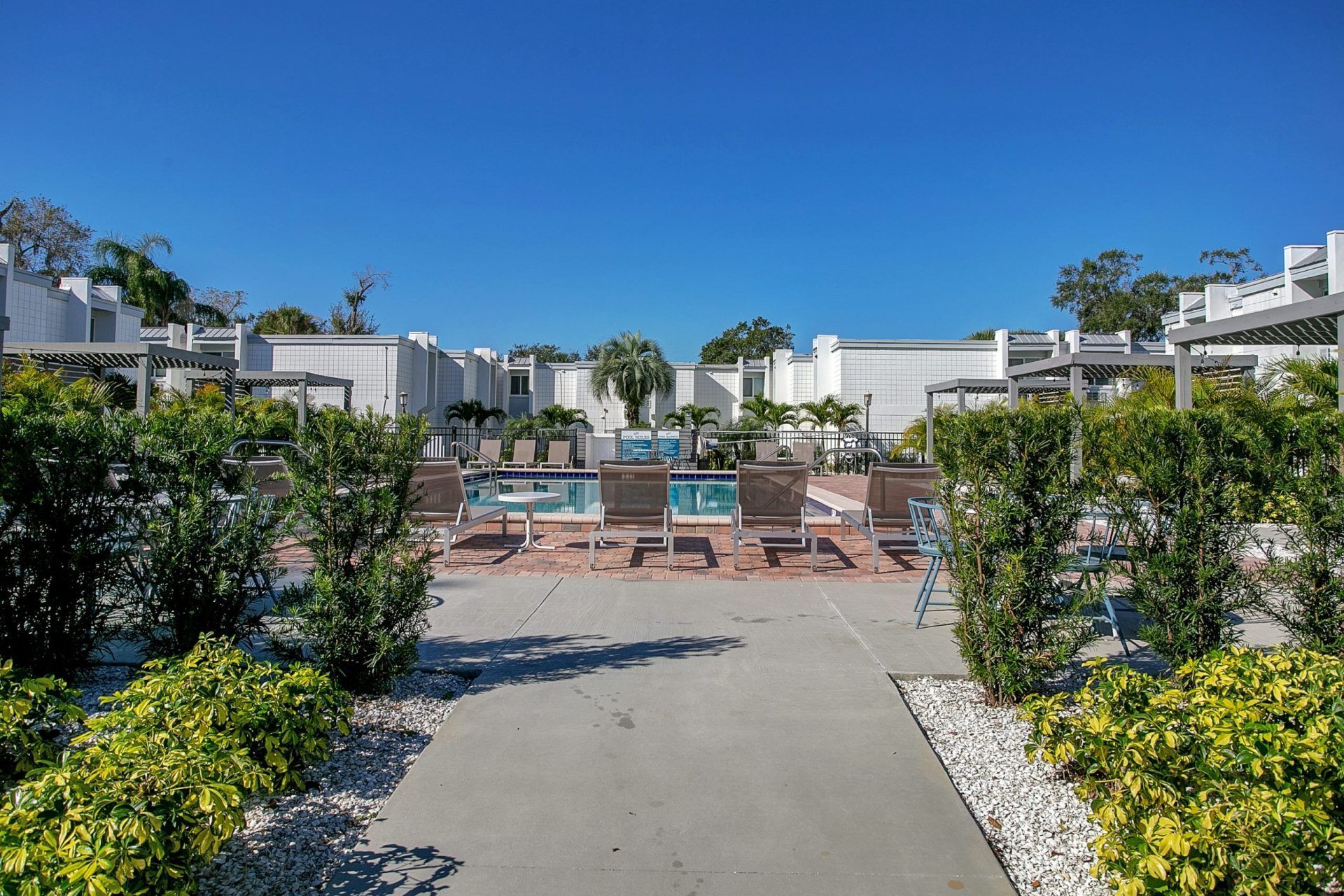 A walkway leading to a large swimming pool surrounded by trees and bushes.
