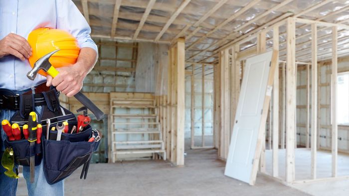A man is holding a hard hat and a tool belt in a room under construction.