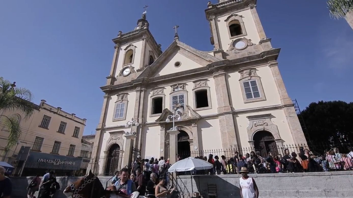 Igreja ornamentada, de cor clara, com duas torres sineiras, com pessoas reunidas do lado de fora em um dia ensolarado.