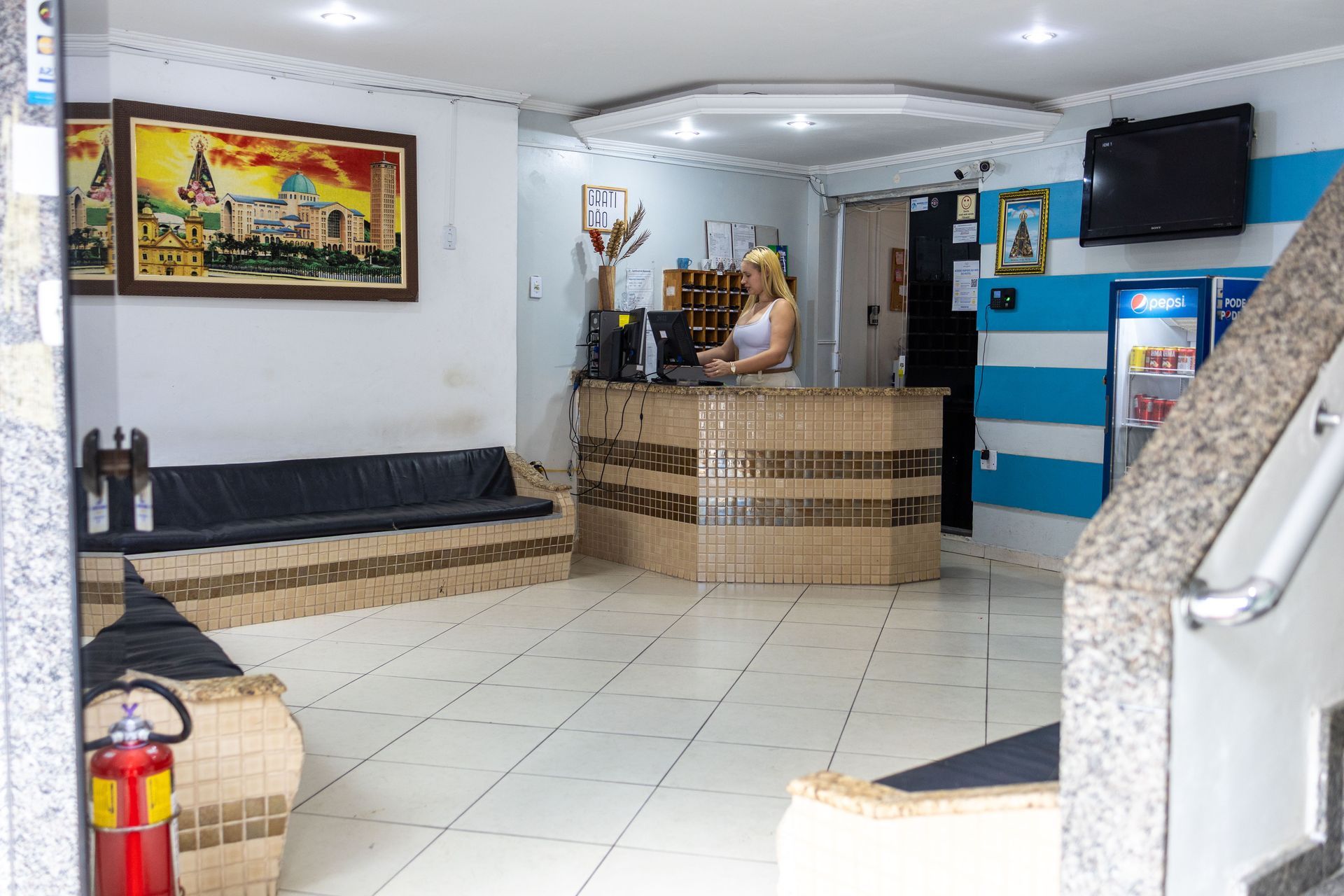 Área de recepção do hotel com uma pessoa ao balcão. Possui piso de azulejo, sofá e obras de arte na parede.