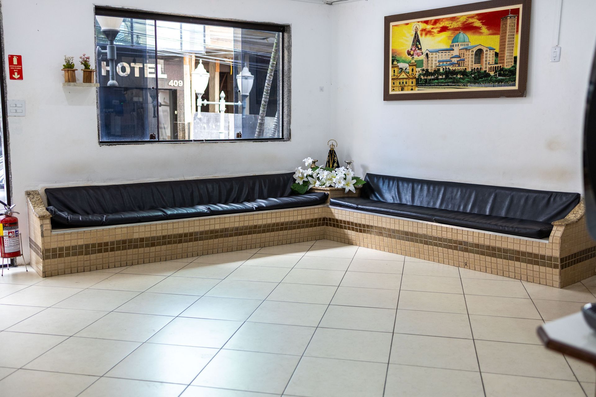 Hall de entrada com piso de azulejo, dois sofás embutidos compridos com almofadas pretas, uma janela, um quadro e um pequeno arranjo de flores.