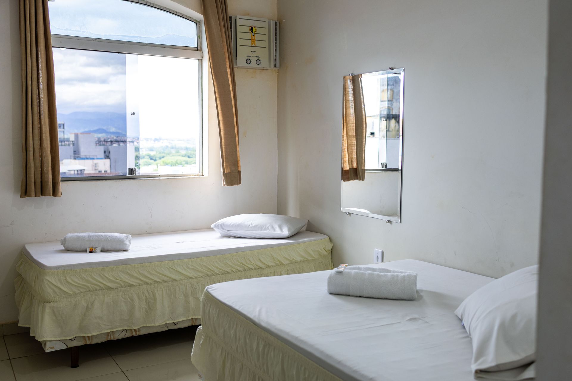 Duas camas de solteiro em um quarto de hotel iluminado, perto de uma janela com cortinas bege. Um pequeno espelho está pendurado na parede.