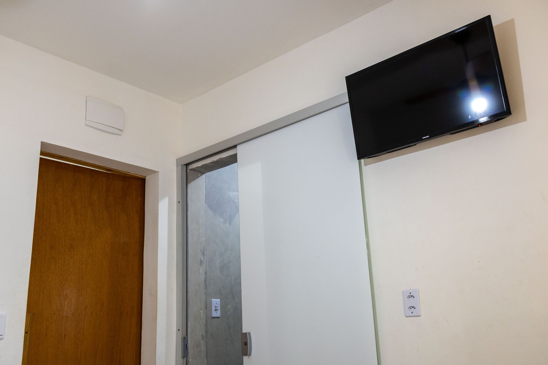 Uma sala com uma TV fixada na parede, uma porta de vidro deslizante e uma porta marrom à esquerda.