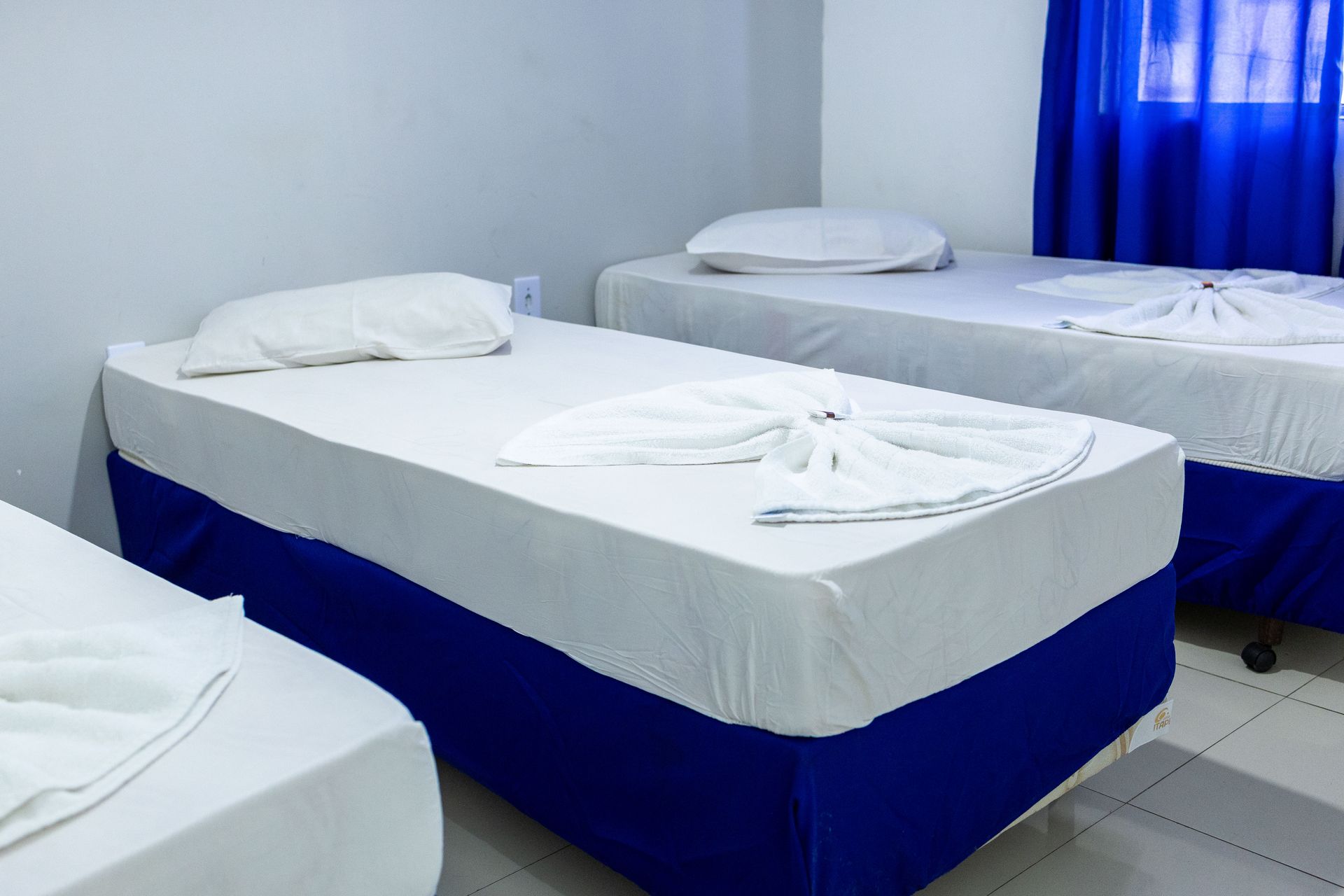 Três camas em um quarto, lençóis brancos, saias de cama azuis, cortinas azuis.