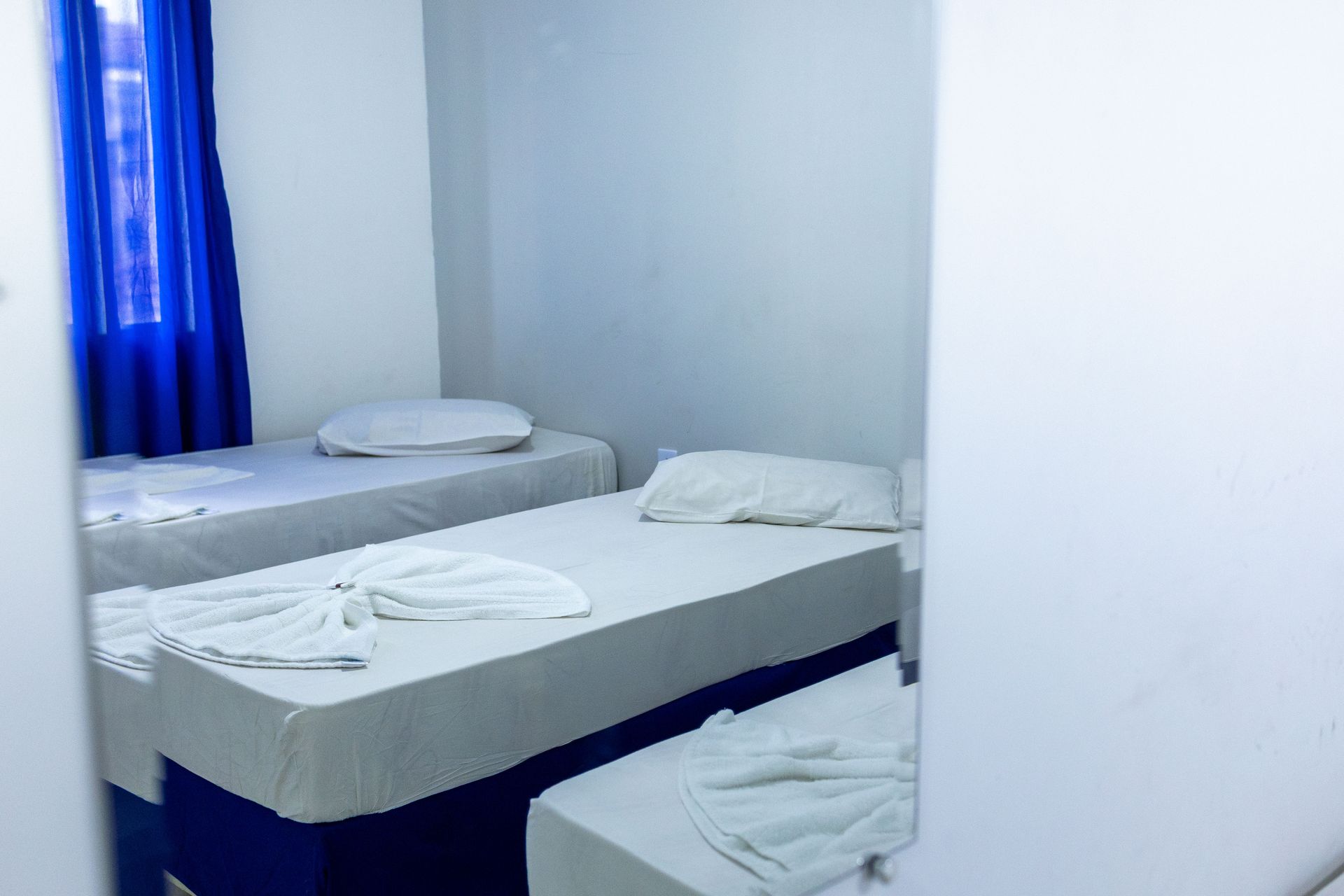 Três camas em um quarto bem iluminado com lençóis brancos, cortinas e detalhes em azul.