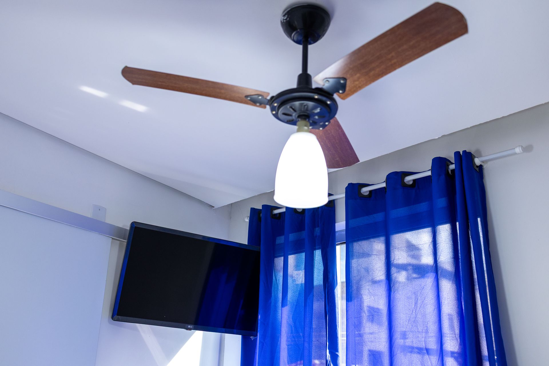 Ventilador de teto com luminária e pás de madeira, acima de uma TV na parede e cortinas azuis.