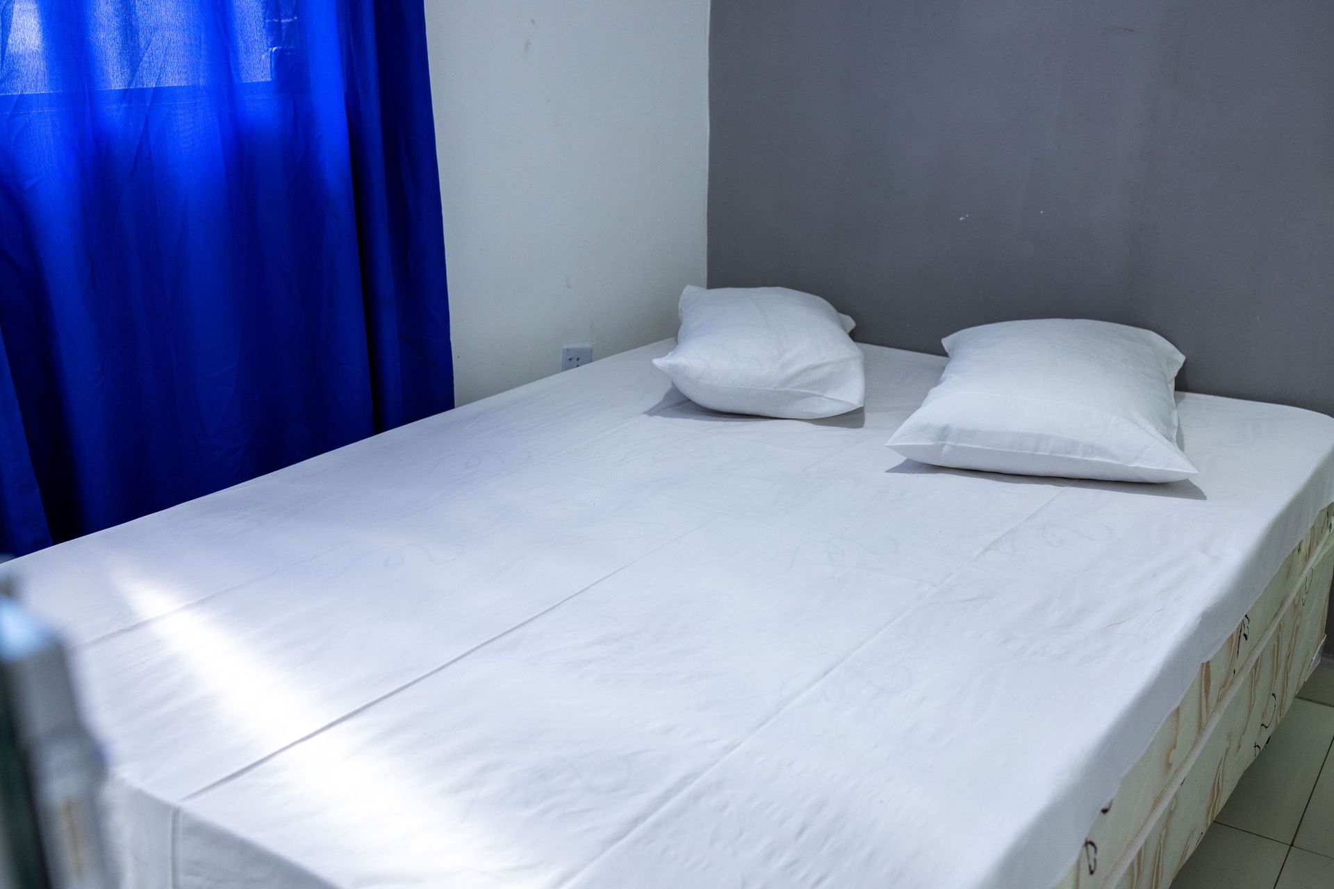 Quarto com cama de casal, roupa de cama branca, dois travesseiros e uma cortina azul.