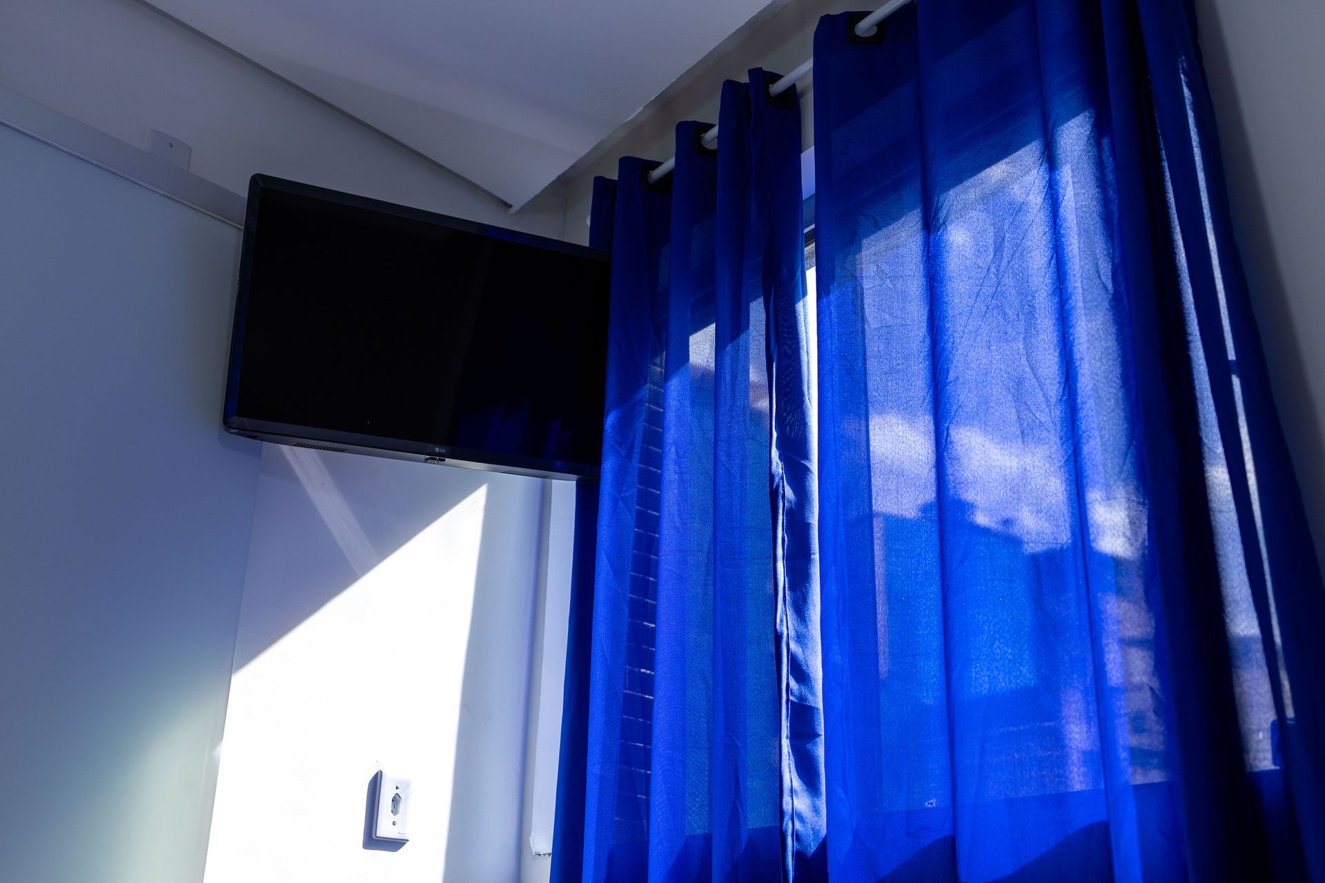 Cortinas azuis ao lado de uma TV fixada na parede. A luz do sol entra pela janela.