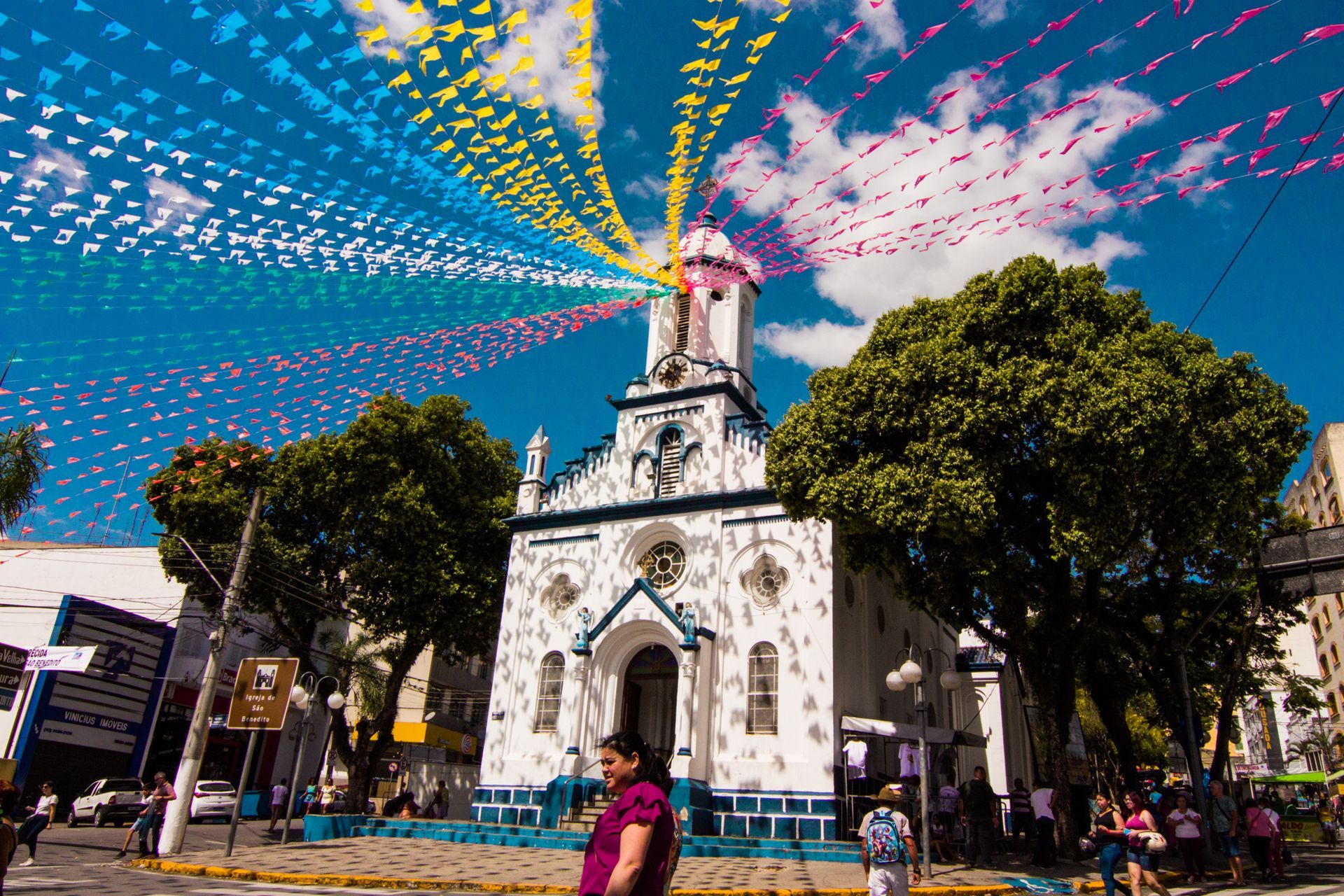 Igreja branca com detalhes em azul, decorada com bandeiras coloridas. Pessoas na calçada sob um céu azul.