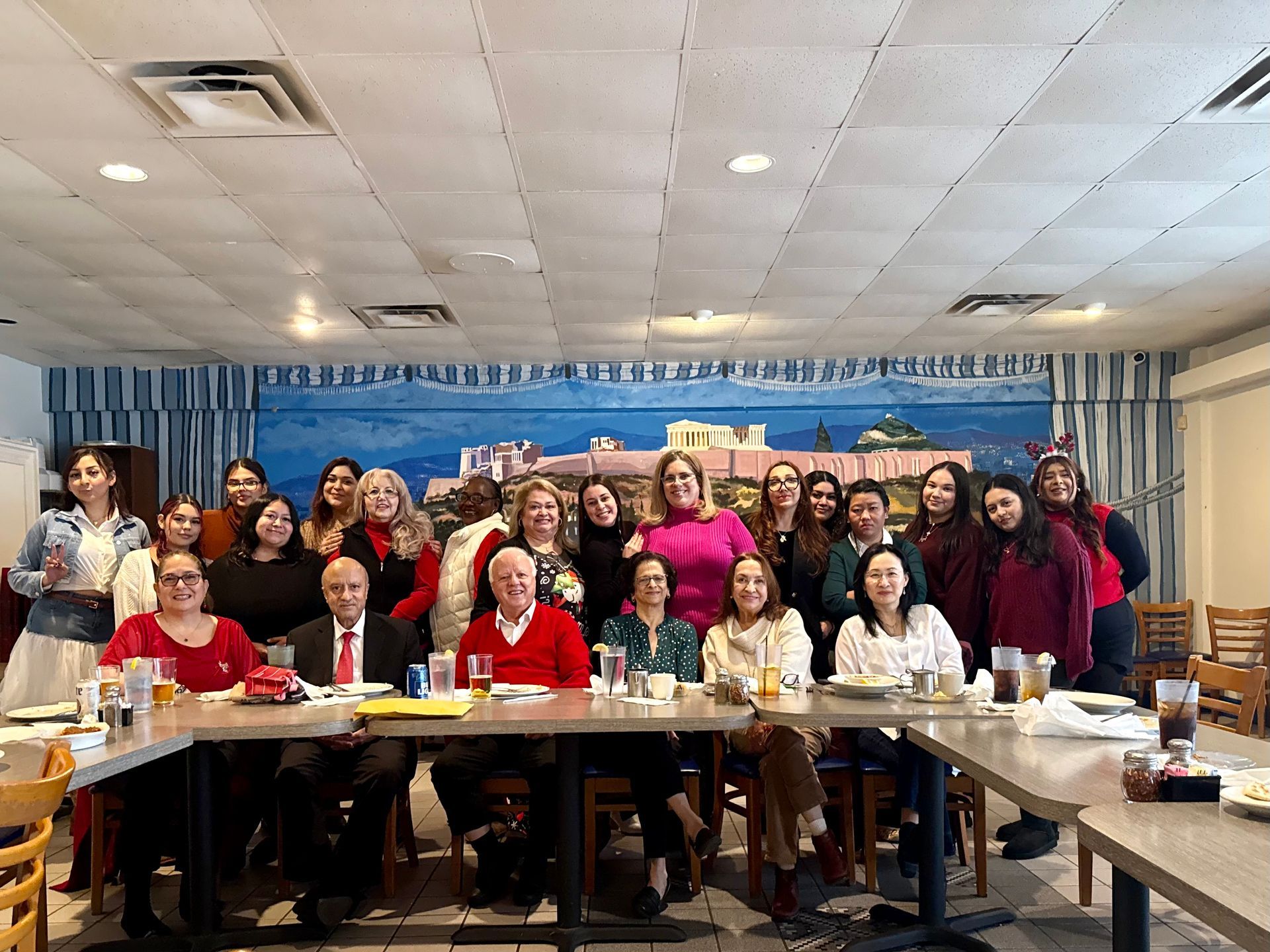 A large group of people smiling at a table during a meeting in a restaurant with a mural of a Greek city on the wall.