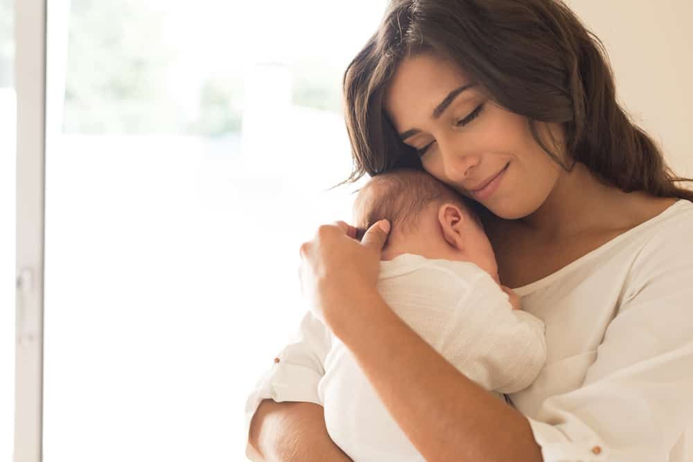 A person with long dark hair smiles gently while cradling a newborn in a white shirt against their chest.