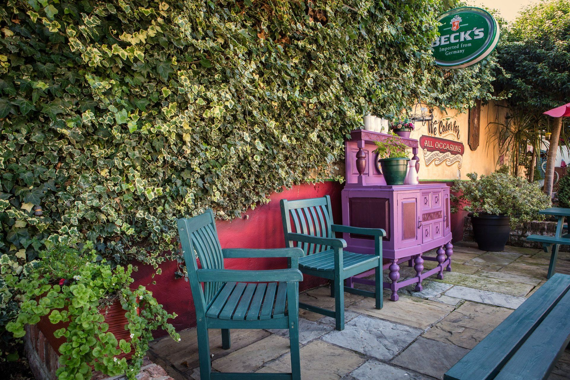 Outdoor seating area with teal chairs, purple cabinet, and green ivy wall. Beck's beer sign.