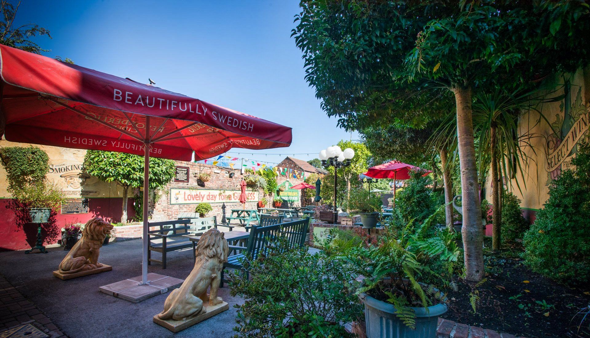 Outdoor restaurant patio with red umbrellas, tables, and lion statues. Lush greenery surrounds the dining area.