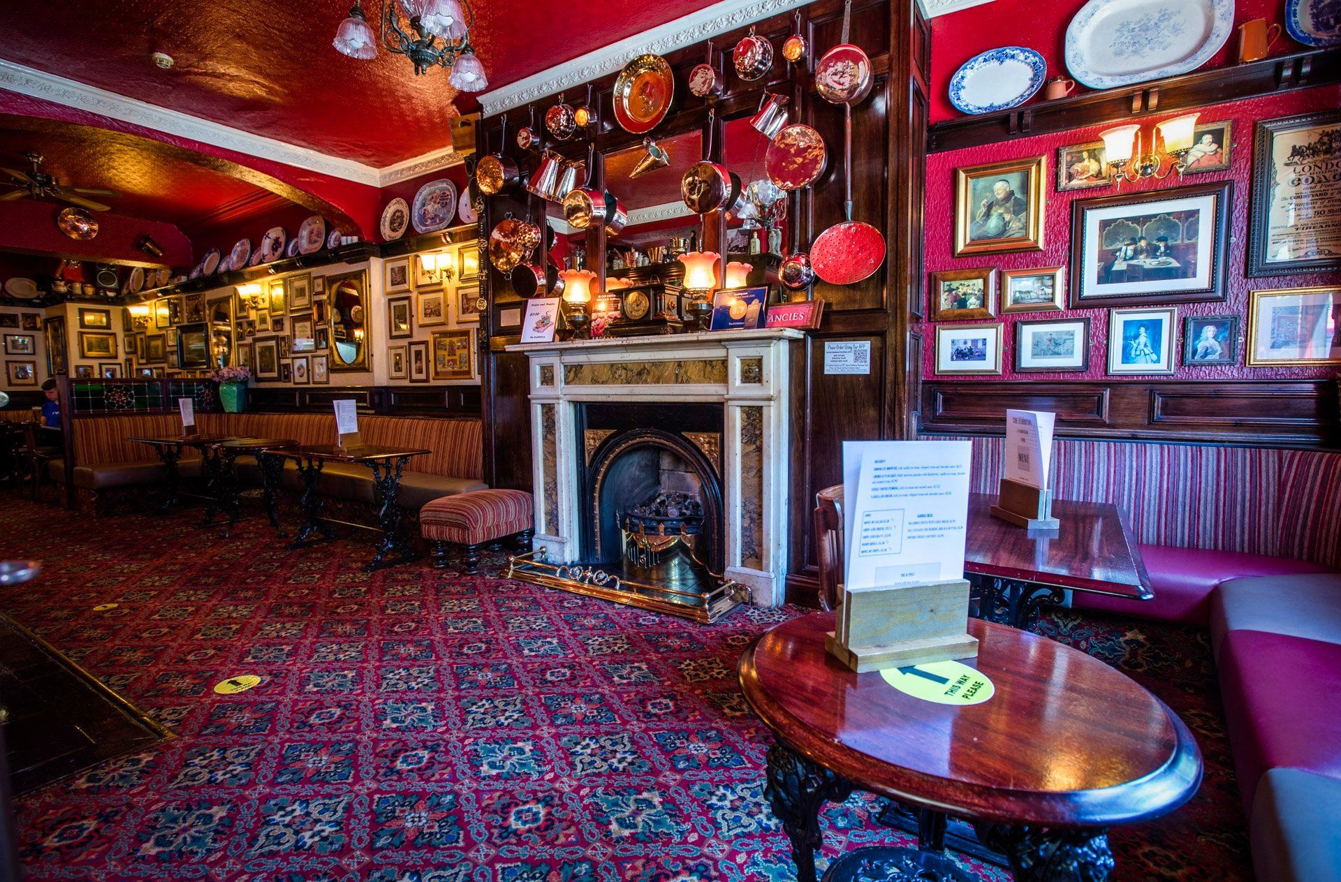 Cozy interior of a pub, red ceiling and carpet. Fireplace, wood paneling, and decor including plates and pictures on walls.