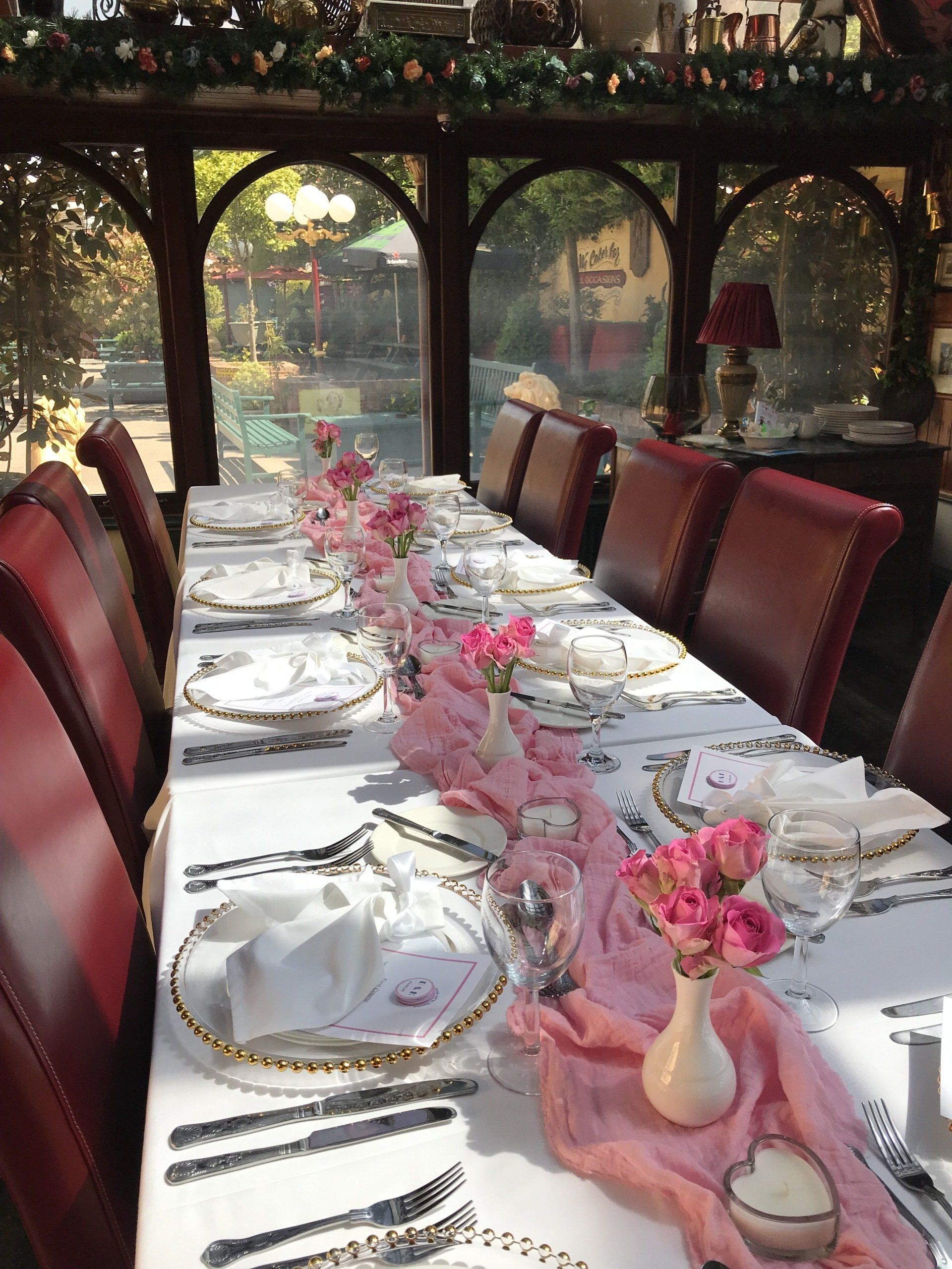 Elegant table setting with pink roses, white tablecloth, and red chairs in a sunroom.