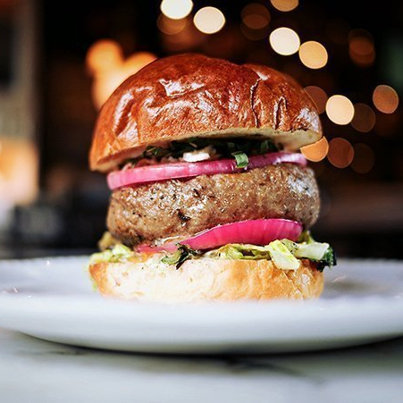 Burger on a bun with red onion, lettuce, and blurred background of lights.