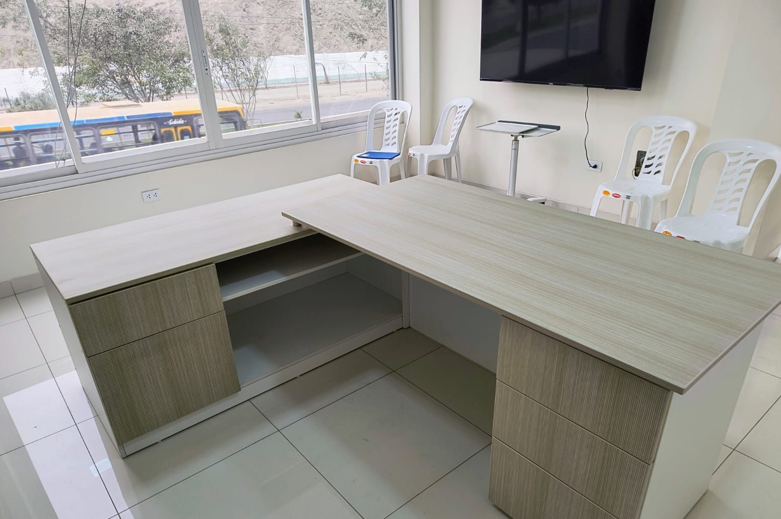 Mostrador de recepción de oficina con mostrador de madera clara en forma de L, sillas y un televisor montado en la pared cerca de una ventana.