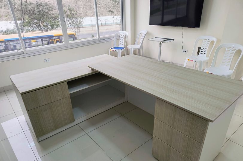 Mostrador de recepción de oficina con mostrador de madera clara en forma de L, sillas y un televisor montado en la pared cerca de una ventana.