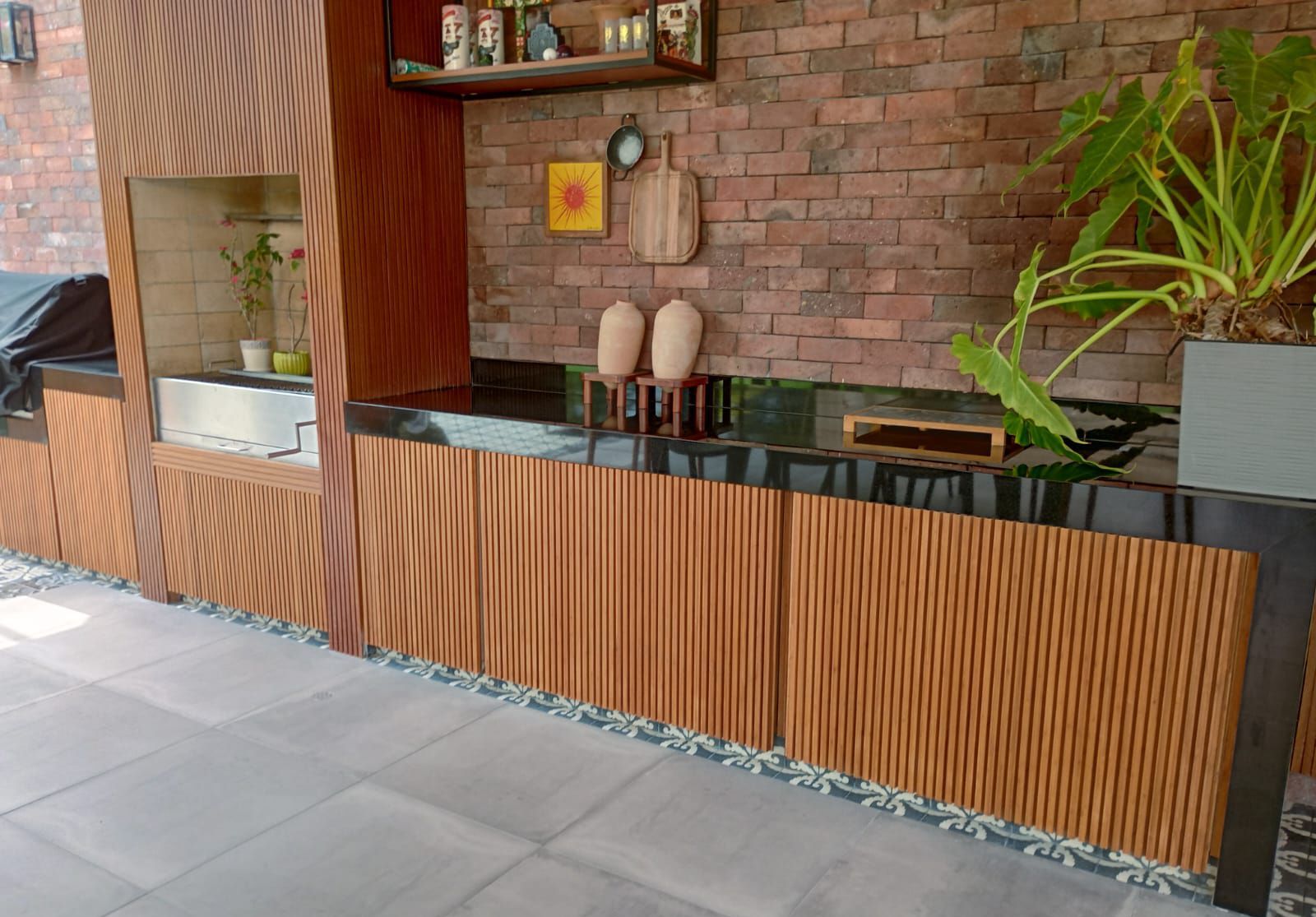 Barra exterior con listones de madera, pared de ladrillo, encimera negra y plantas en macetas.