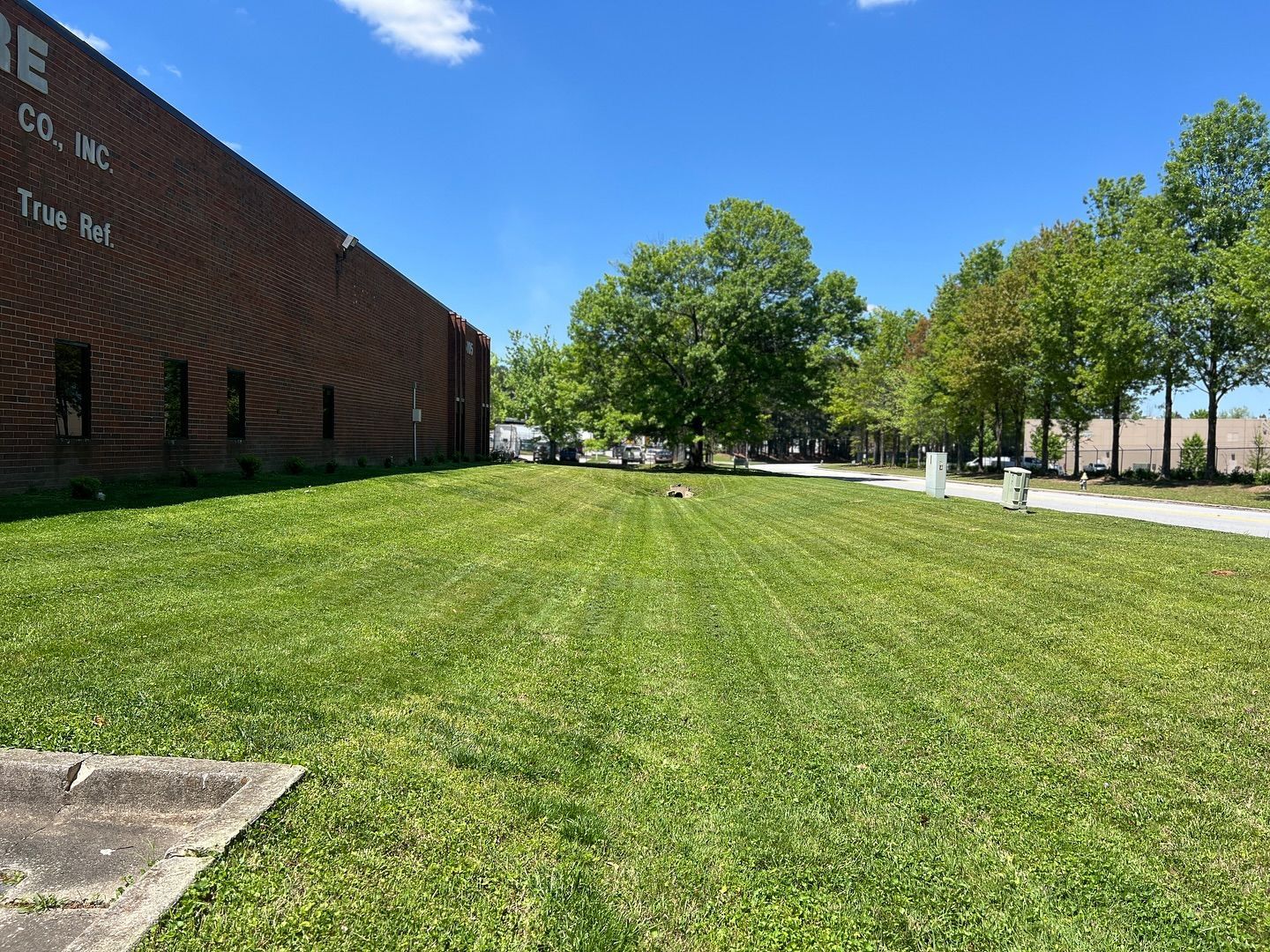 Lush green lawn with a brick building on the left and trees lining a path on the right under a blue sky.