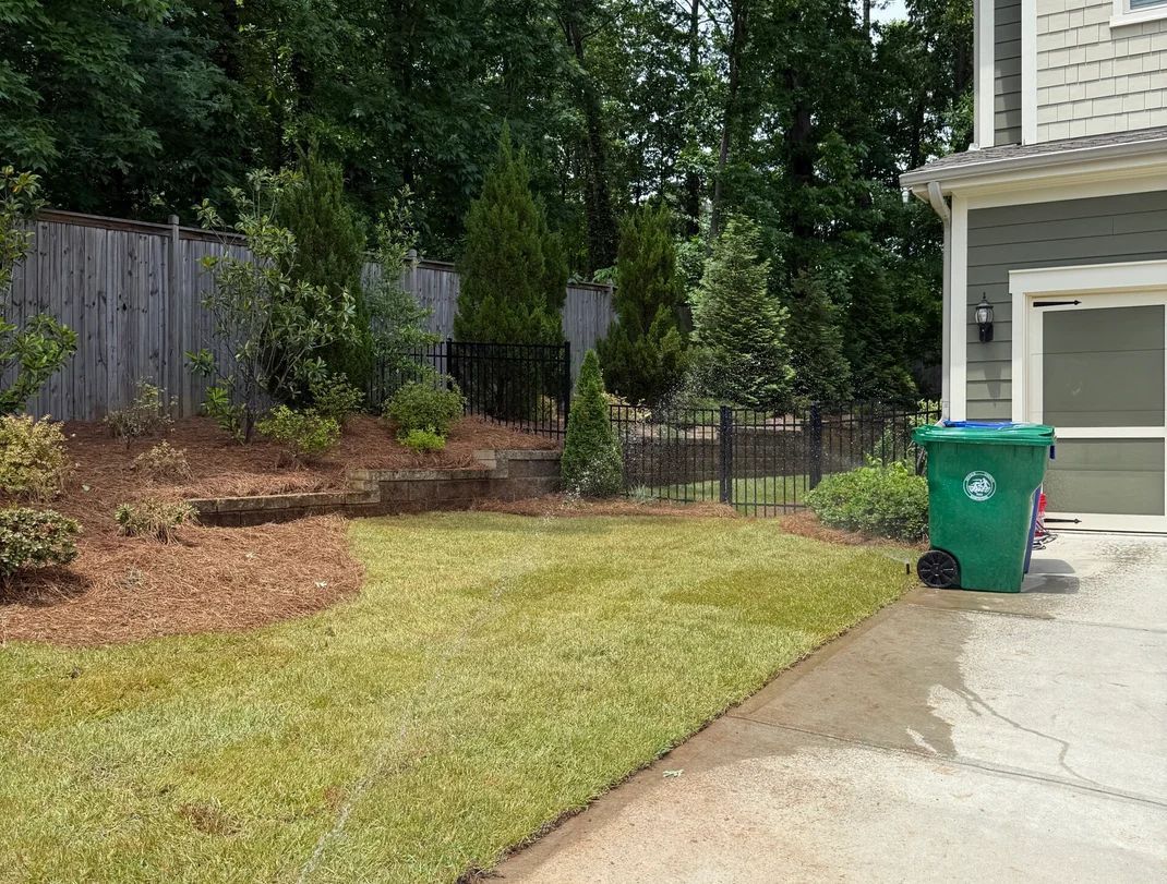 Lawn with green grass, a green trash bin, fence, and trees, next to a house with a door.