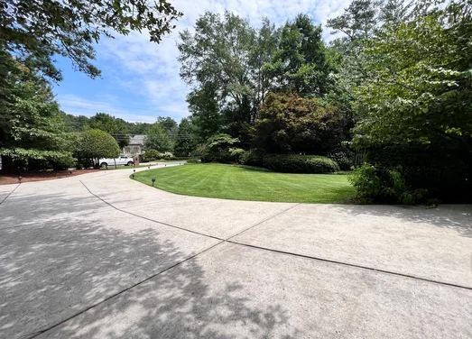 Concrete driveway curving towards a green lawn surrounded by trees and foliage under a blue sky.