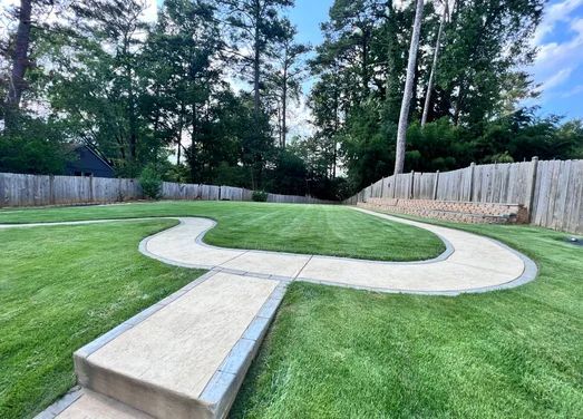 Concrete path winding through a green lawn in a backyard with trees and a wooden fence.