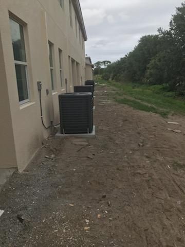 A row of air conditioning units outside a tan building on a dirt path, with trees in the background.