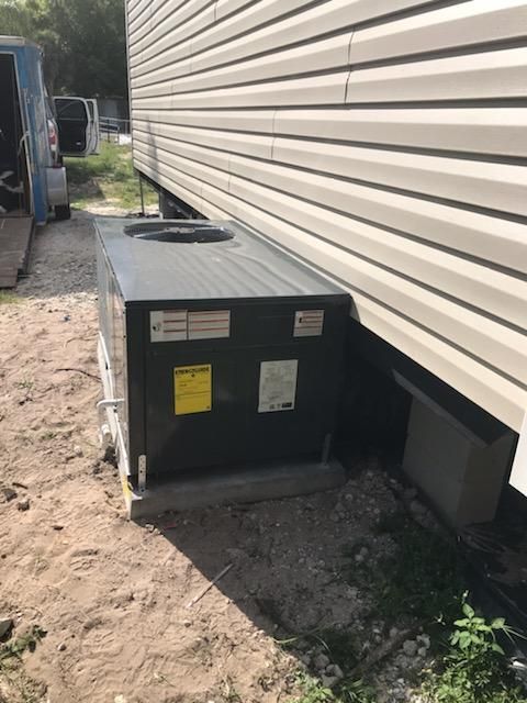 HVAC unit next to a house. Gray siding, concrete base, and sandy ground.