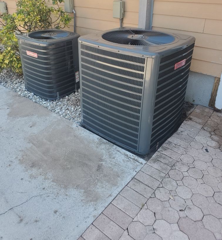 Two Goodman air conditioning units on a concrete slab next to a house; one unit is larger.