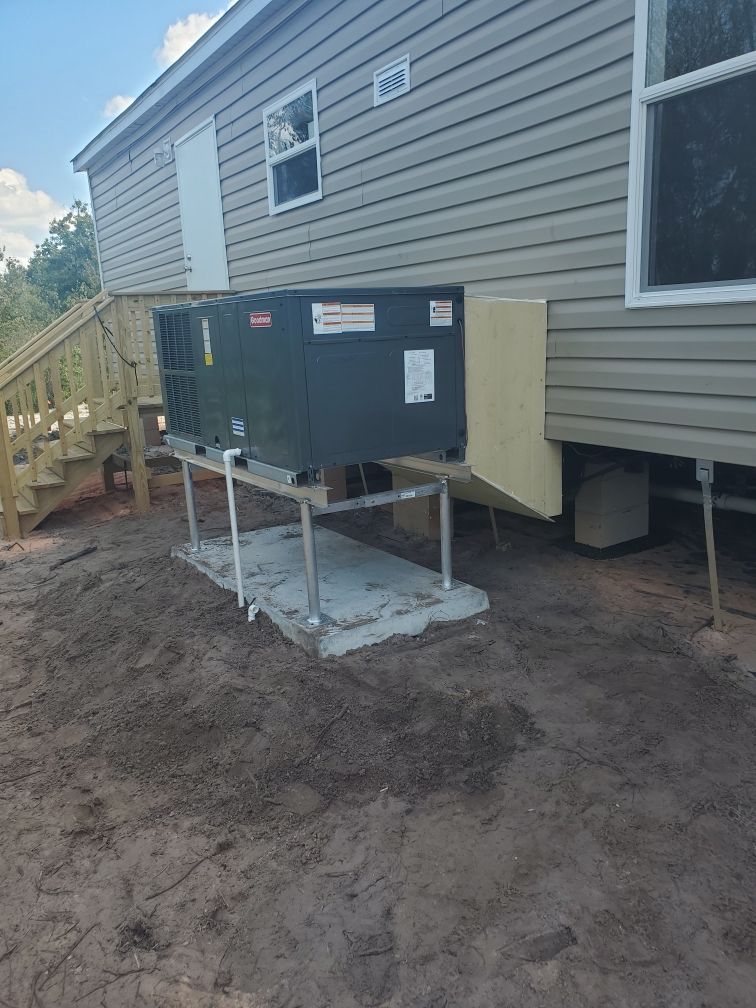 A dark HVAC unit sits on a raised metal platform next to a mobile home on dirt ground.