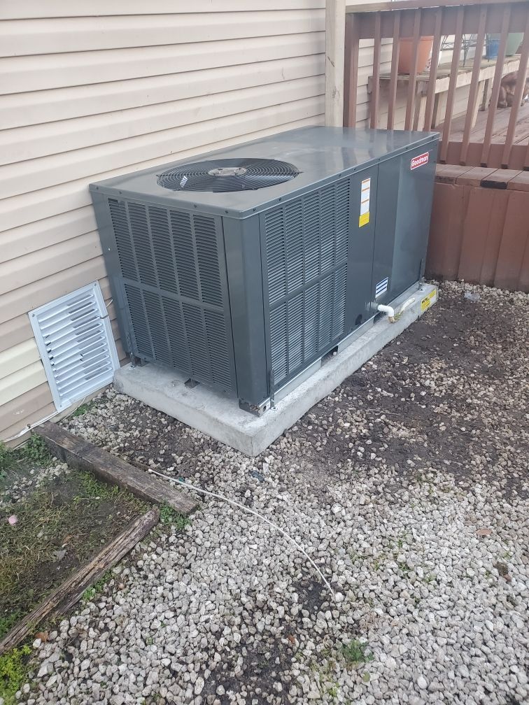 A dark gray Bryant air conditioning unit sits on a concrete pad next to a house with beige siding and a wooden deck.