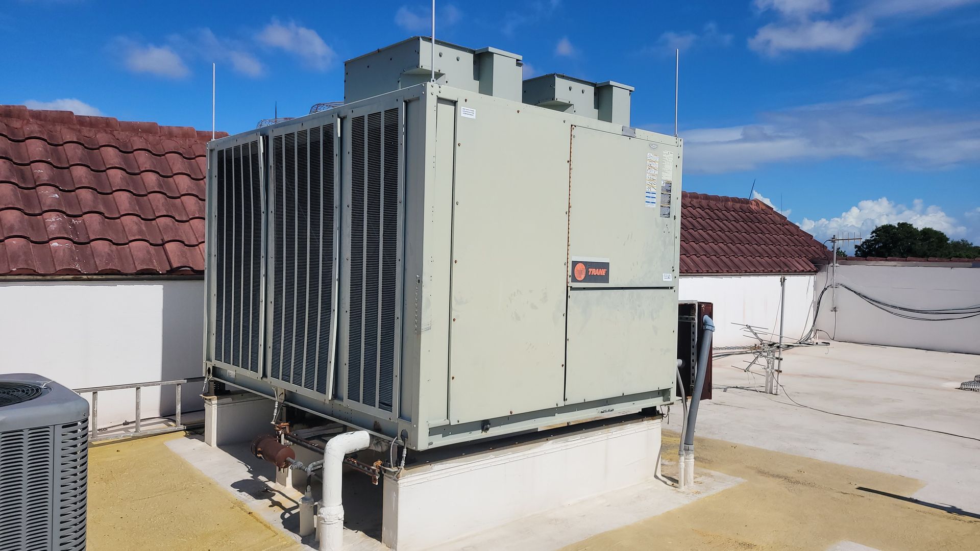 A large Trane rooftop HVAC unit on a flat roof with red-tiled roofs in the background under a blue sky.