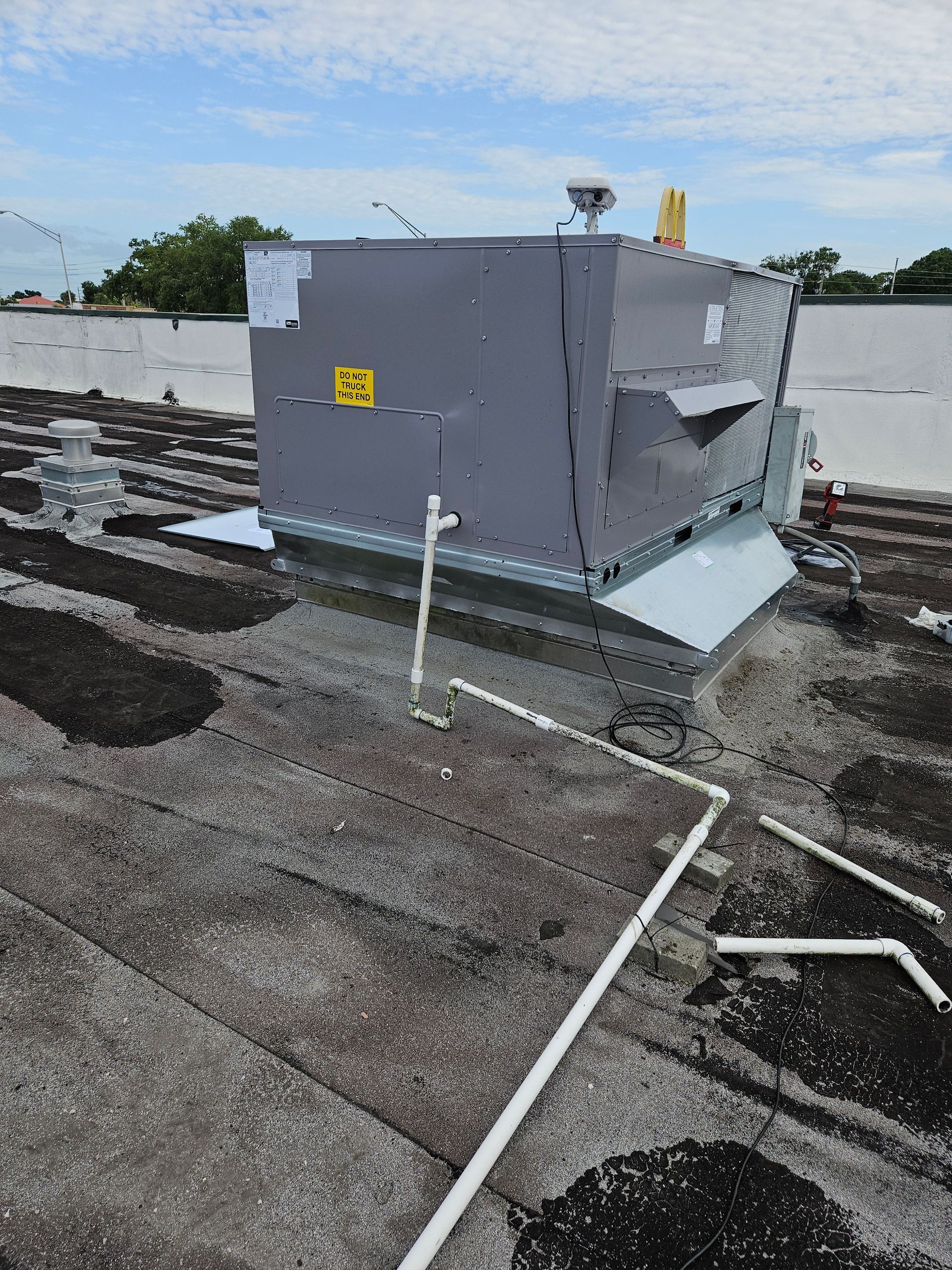 Rooftop HVAC unit on a flat, dark roof with white pipes and a blue sky.