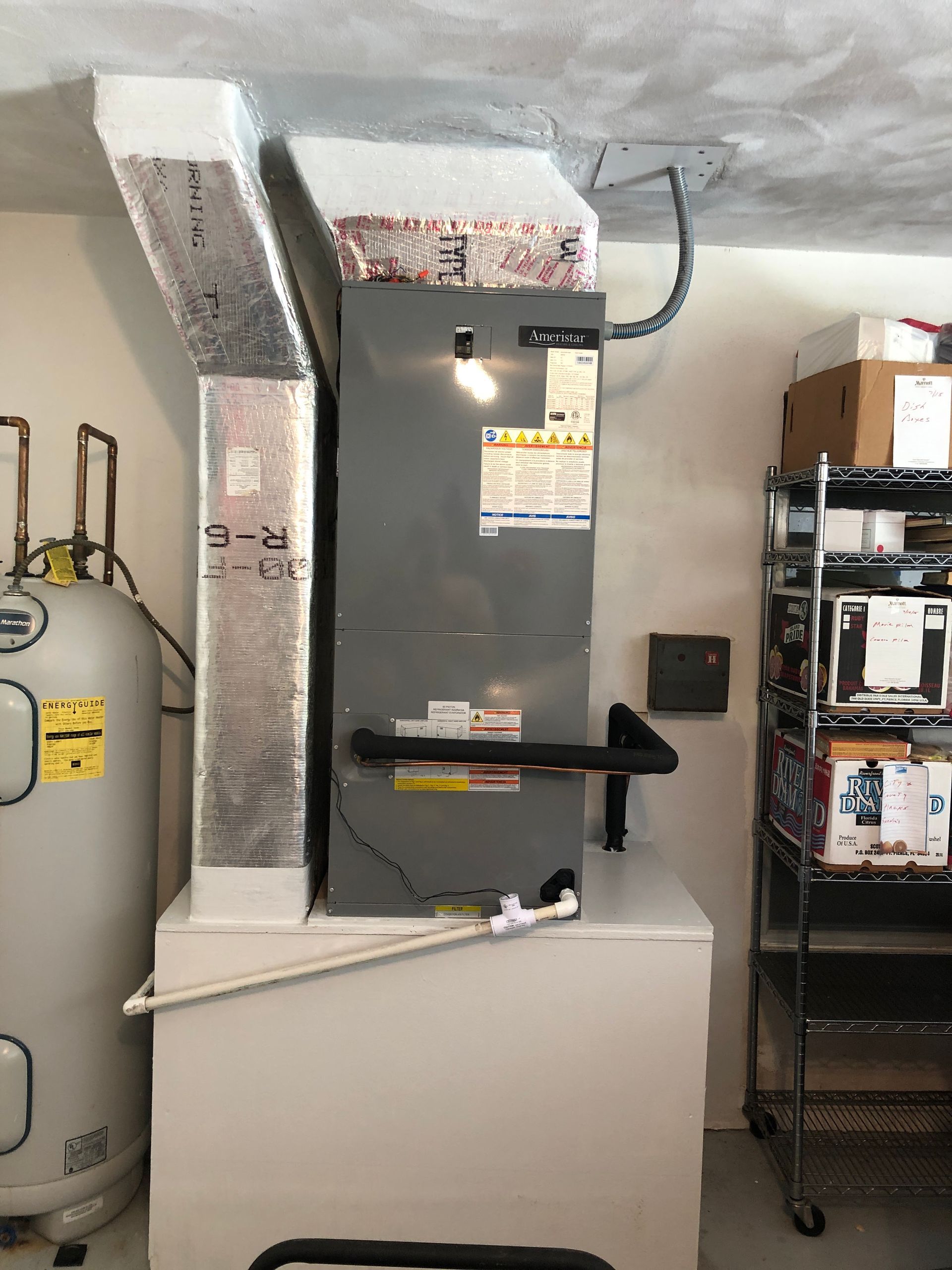 A gray HVAC unit on a white base in a garage setting with a water heater and shelving unit.
