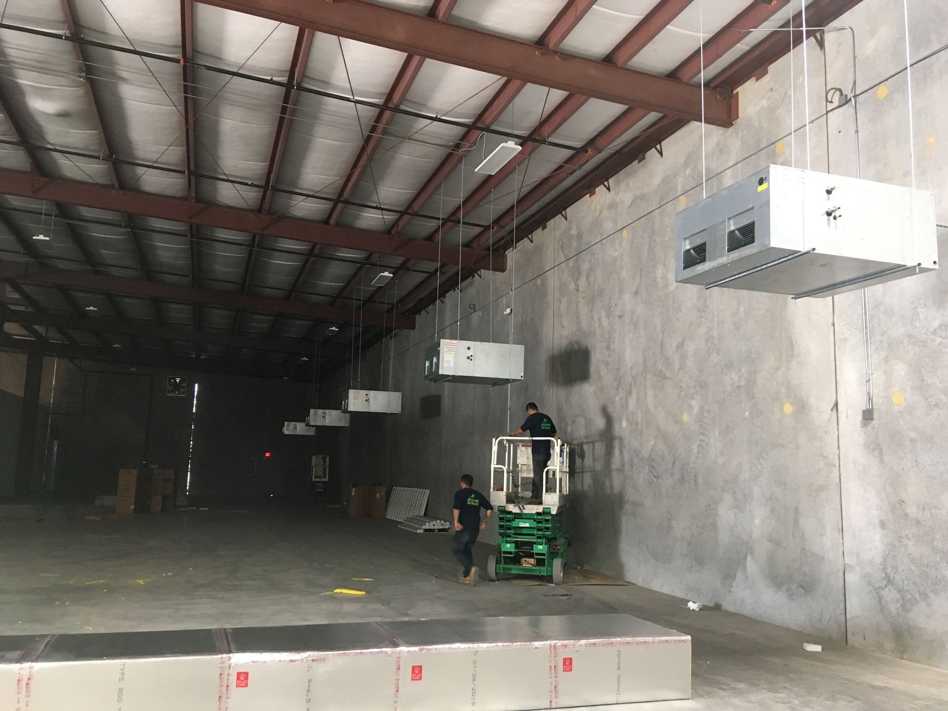 Inside a warehouse, workers on a lift install HVAC units suspended from the ceiling.