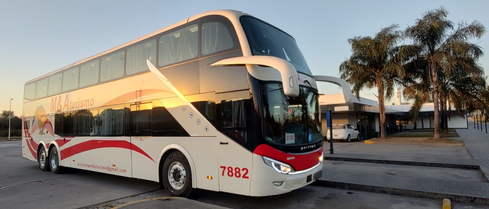Autobús de dos pisos estacionado en una terminal, con un diseño rojo y blanco; palmeras en el fondo.