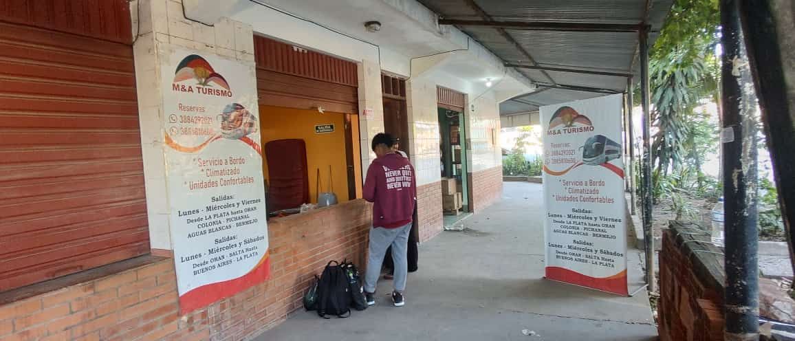 Persona parada frente a una tienda con pancartas. La tienda tiene una ventana abierta y una pared amarilla visible.