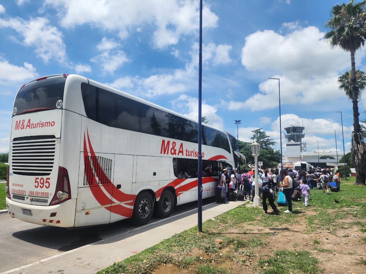 Autobús blanco de M&A Turismo con pasajeros, estacionado cerca de un edificio y gente. Cielo azul y palmeras.