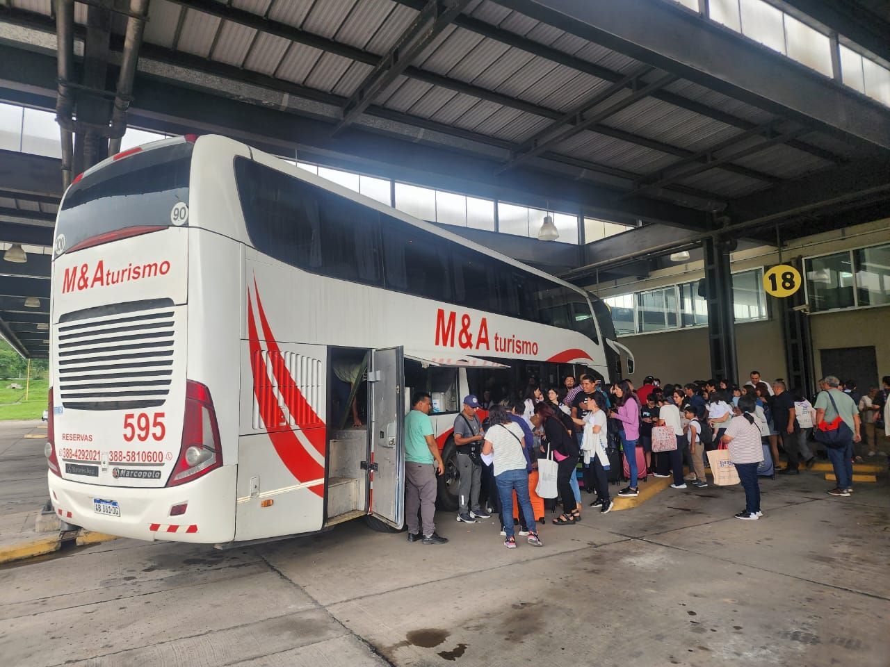 Autobús con pasajeros subiendo a una terminal. Autobús blanco con detalles en rojo, gente esperando en fila.
