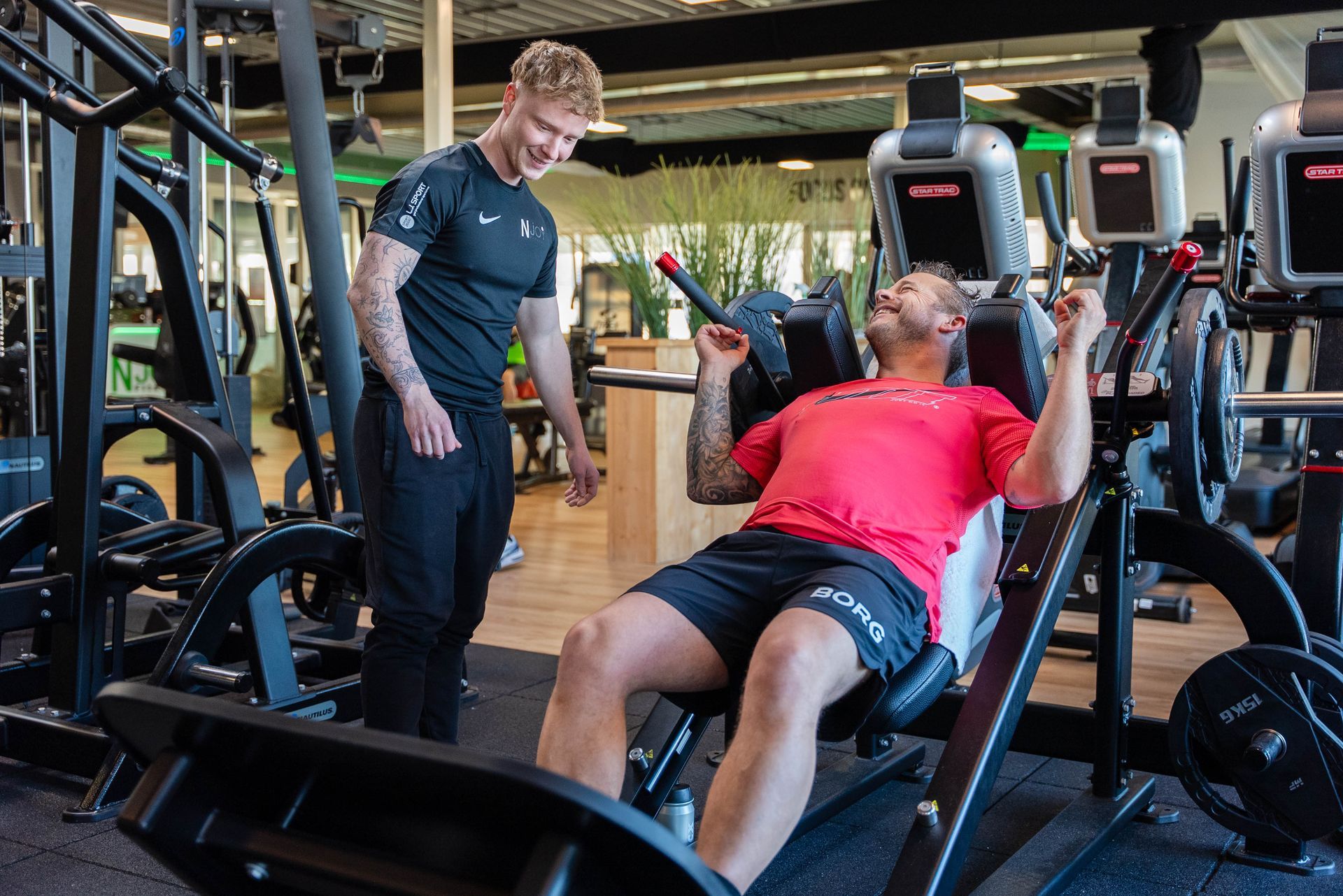 Een man gebruikt een legpress-apparaat in een sportschool met hulp van een trainer. Beiden glimlachen.