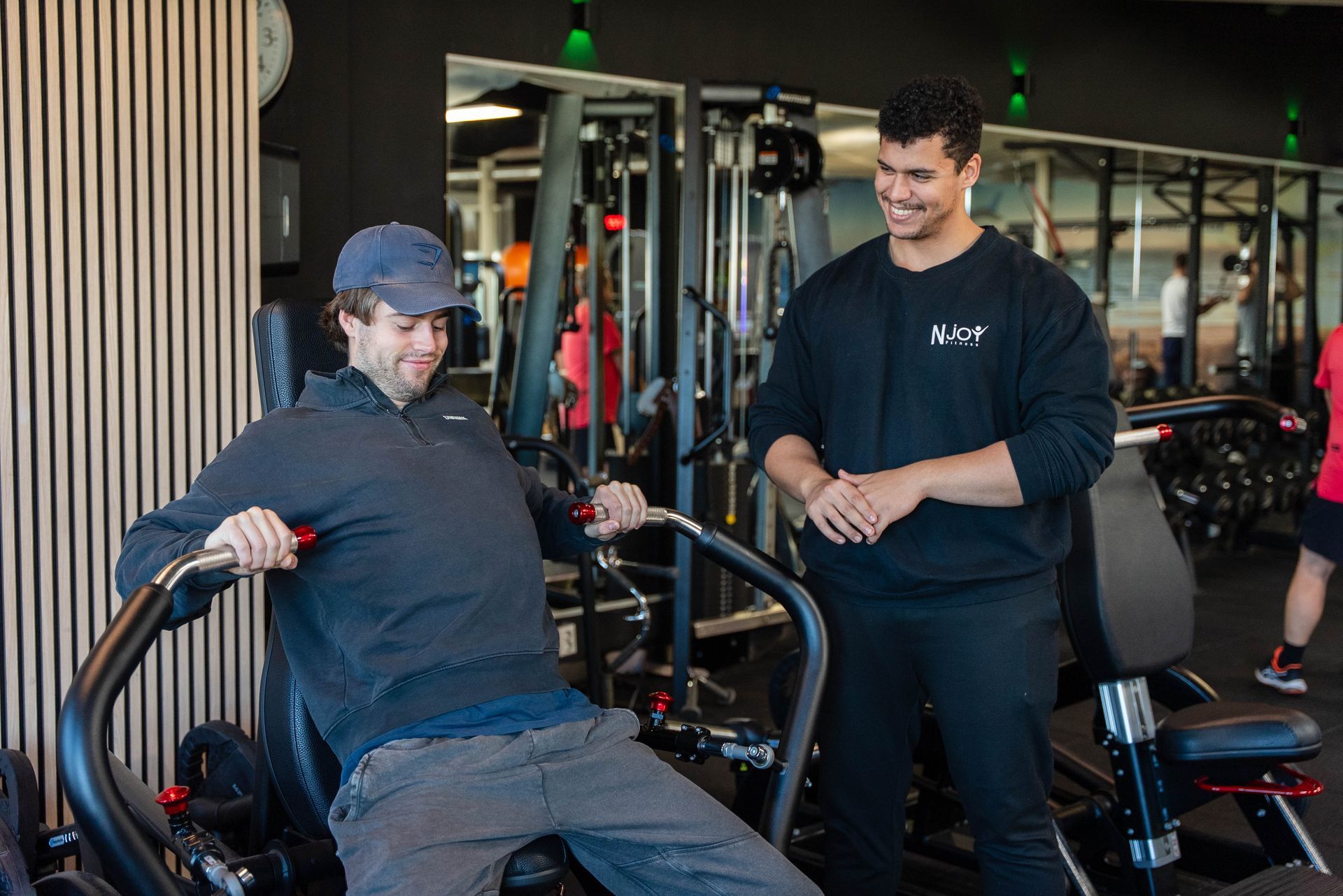 Een man gebruikt fitnessapparatuur, lachend, terwijl een trainer toekijkt. Sportschoolomgeving.