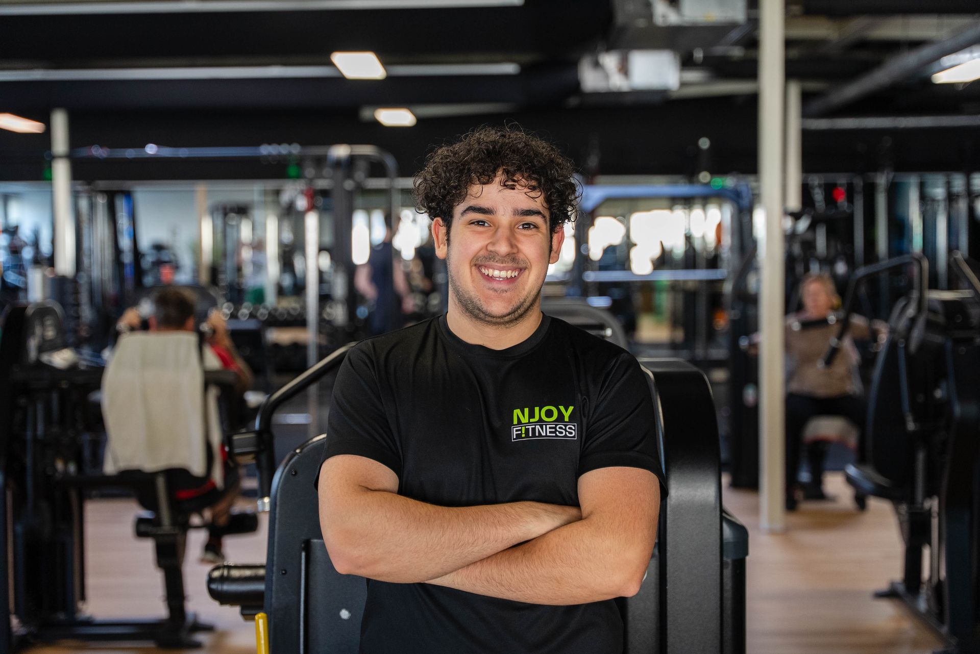 Een man met krullend haar en gekruiste armen lacht in de sportschool, omringd door fitnessapparatuur.