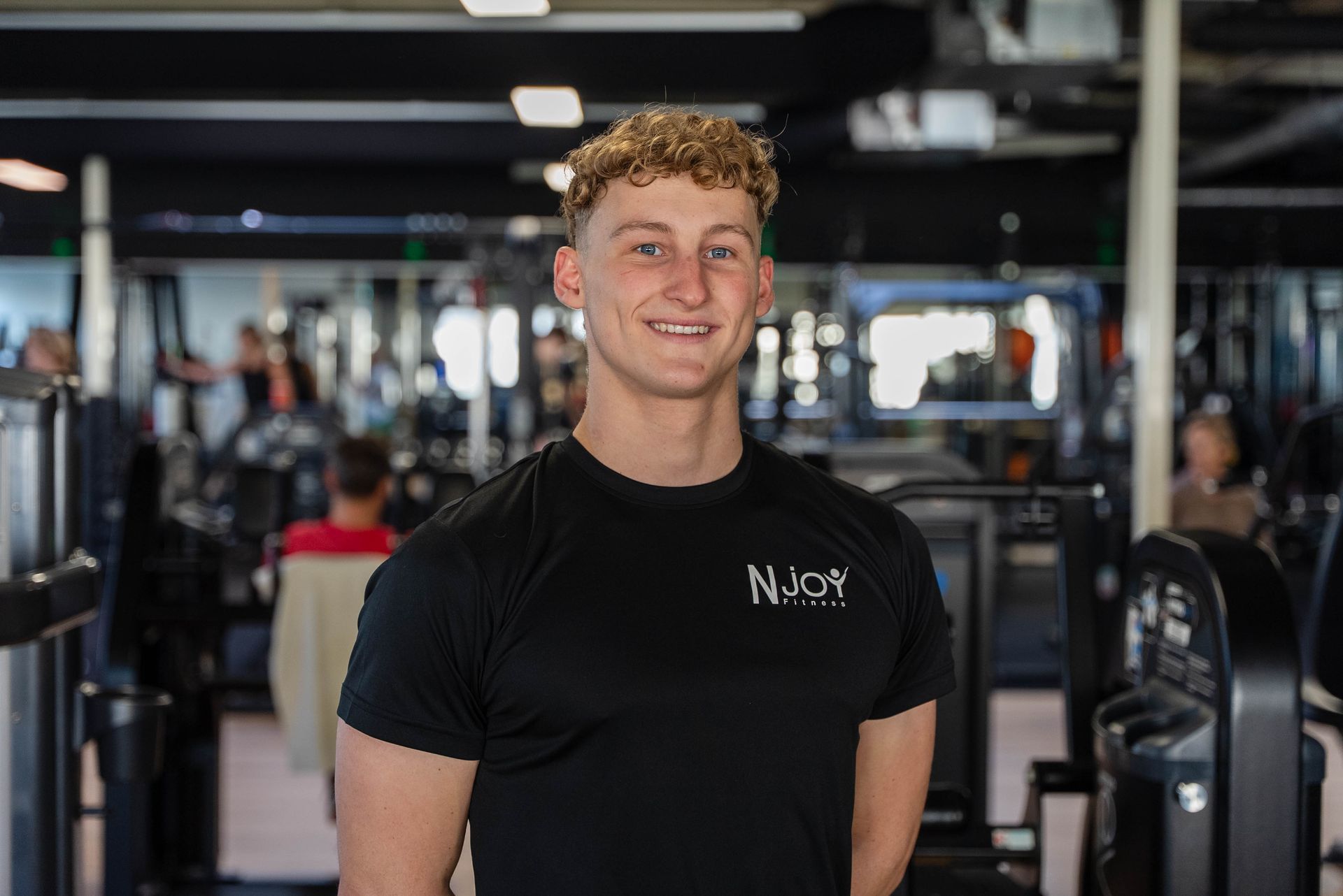 Een man lacht in een sportschool, draagt ​​een zwart shirt met logo en is omringd door fitnessapparatuur.