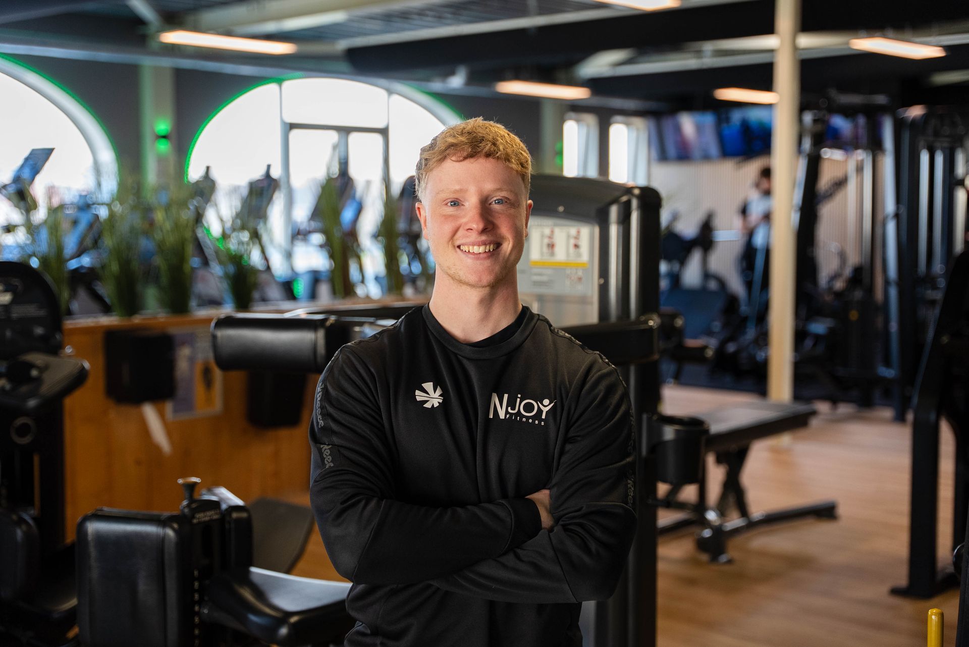Man in zwart shirt met gekruiste armen, lachend in een sportschool.