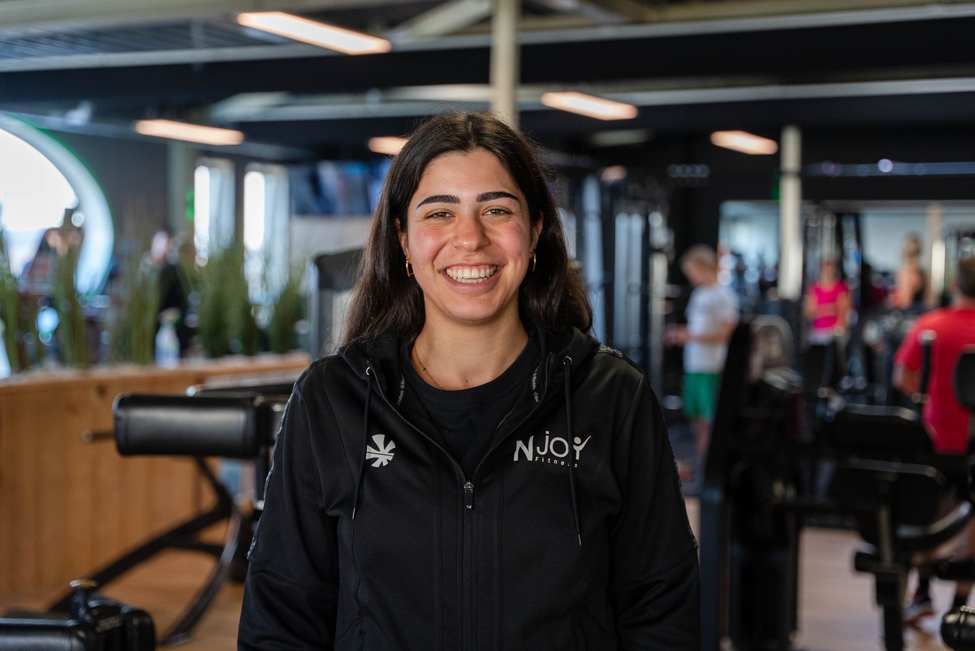 Een vrouw in een zwarte jas lacht in een sportschool, met fitnessapparatuur en mensen op de achtergrond.