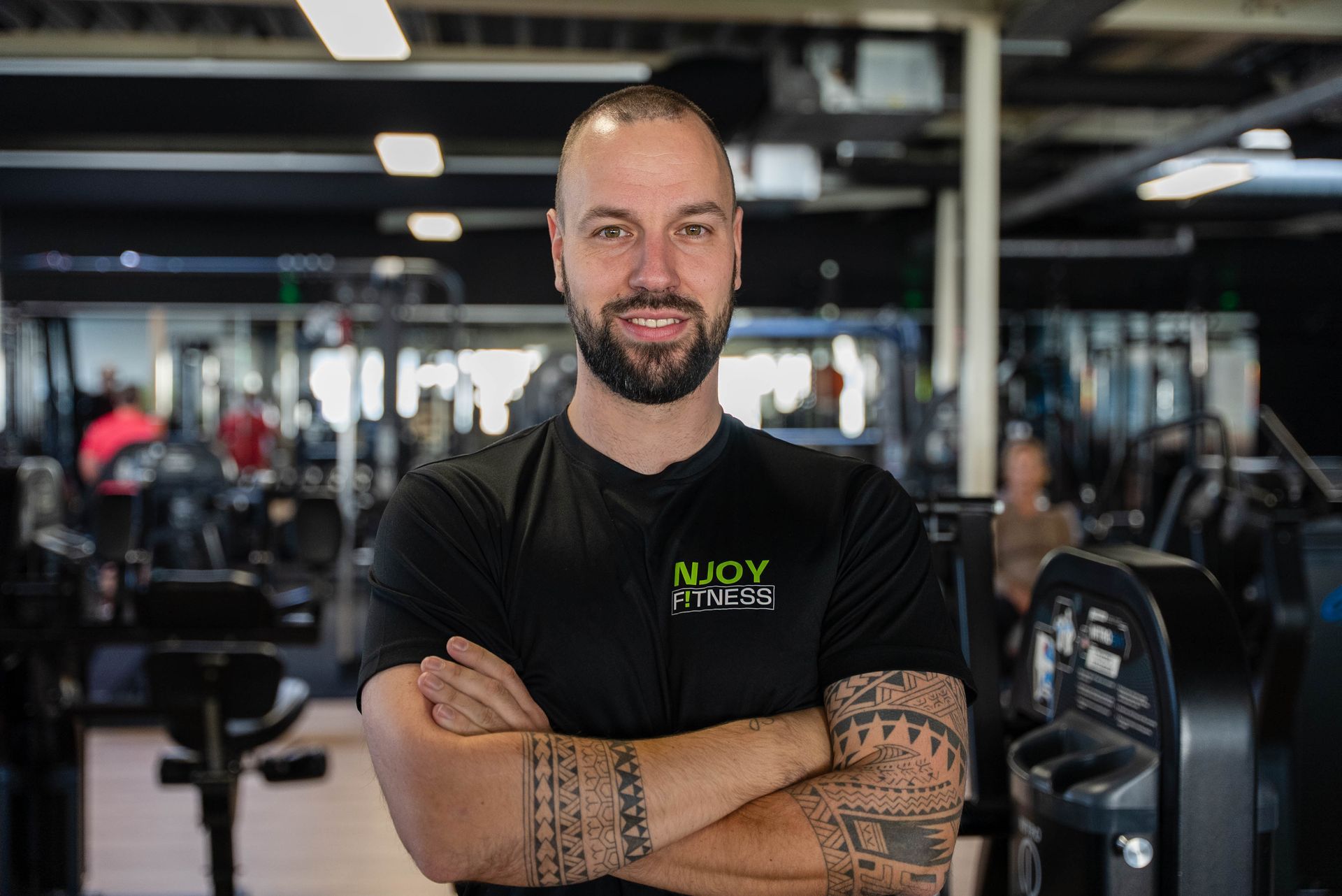 Een man met tatoeages, met de armen over elkaar, lachend in een sportschool.
